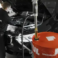 Mechanik wymienia olej w czarnym BMW. Obok stoi duża, pomarańczowa beczka z olejem. Pamiętaj, co ile wymiana oleju jest kluczowa dla silnika.