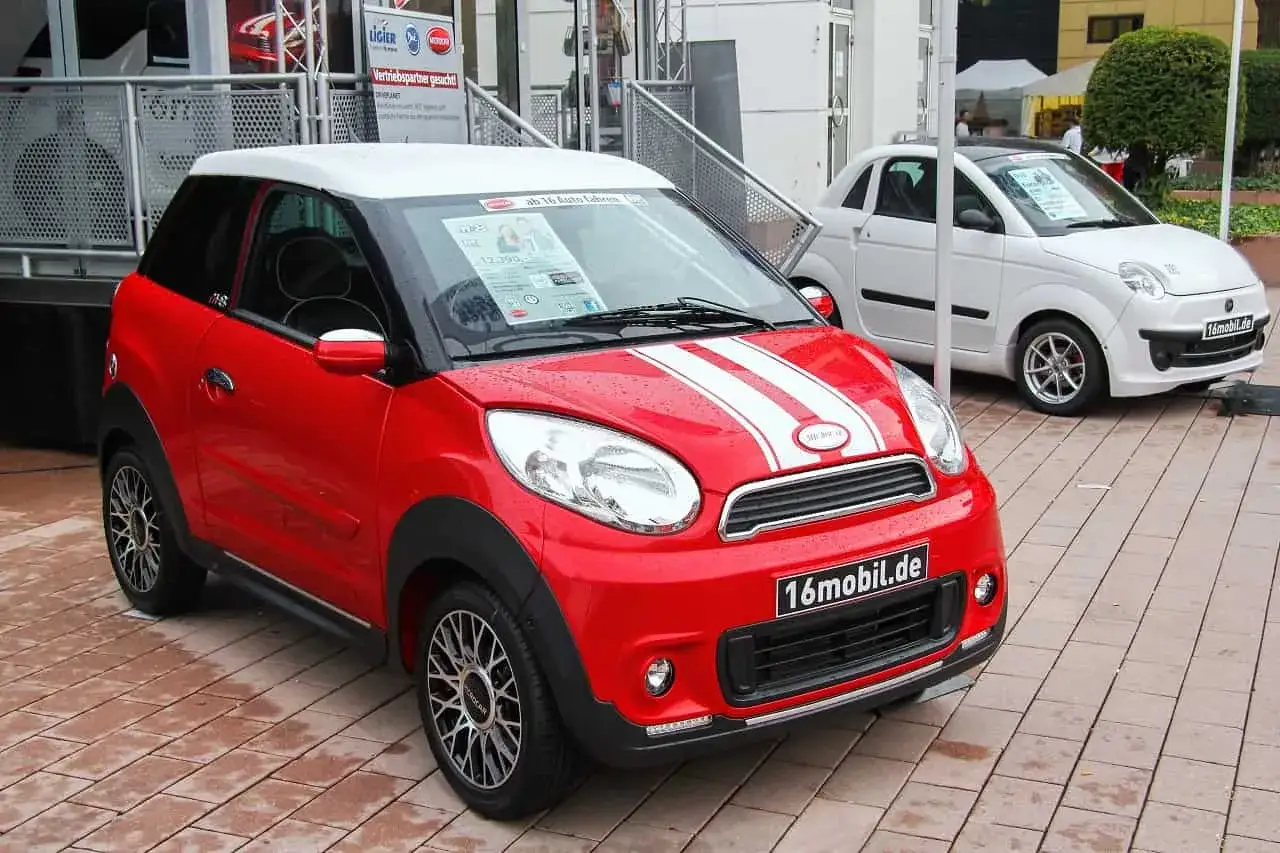 Czerwony samochód bez prawa jazdy, model Microcar, z białymi pasami na masce, obok białego auta. Idealny dla młodych kierowców.